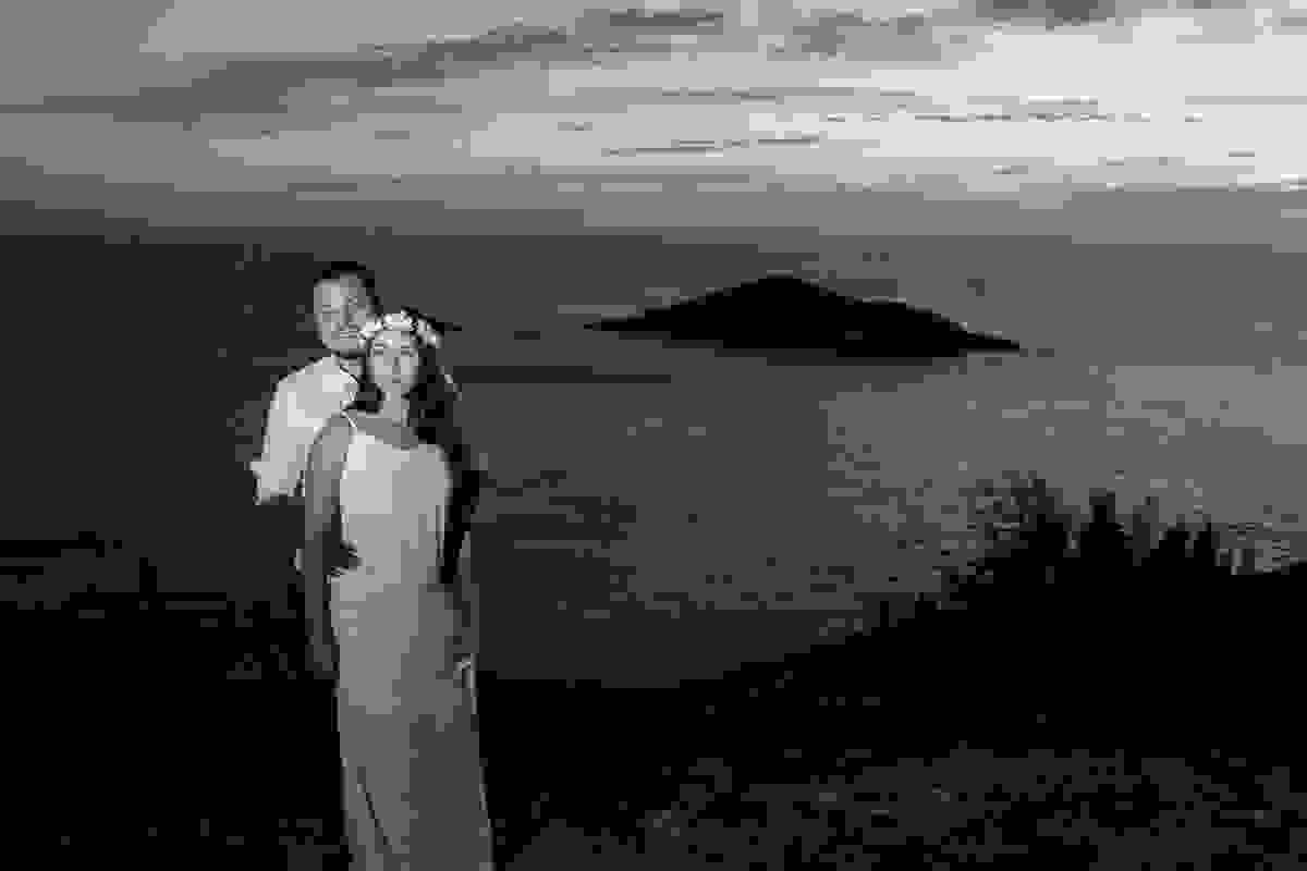 Fotografia no alto do atalaia em arraial do cabo mostra casal de pé sorrindo com o lindo por do sol atraz