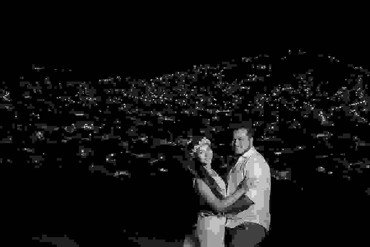 Fotografia noturna do alto do atalaia mostras as casas ilumidas com suas pequenas luzes e casal sorrindo olhando para camera na ultima foto do ensaio pré casamento