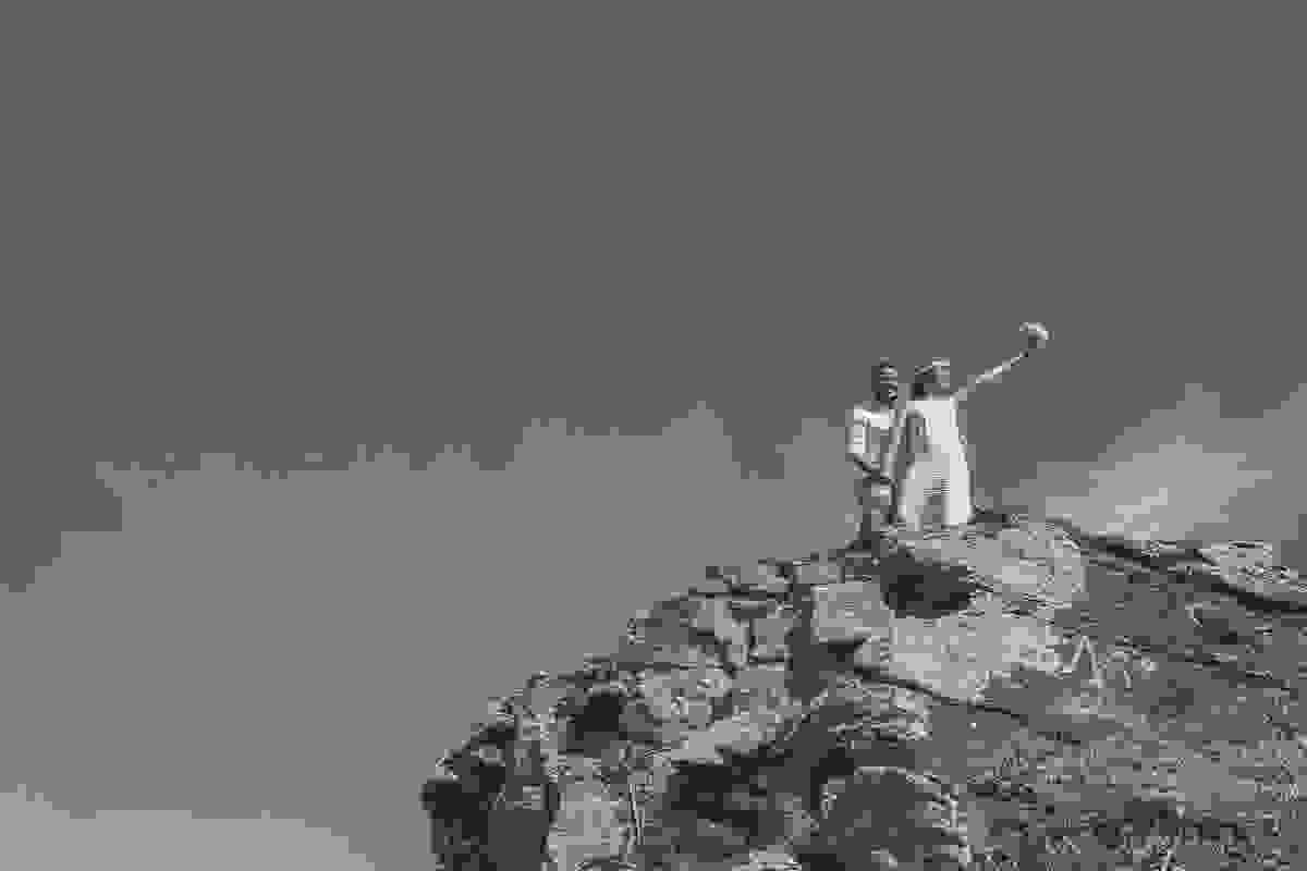 casal bem no alto de uma pedra a noiva segura o bouquet com os braços esticados enguanto noiva a observa em fotografia de pré casamento na praia de cabo frio rio de janeiro