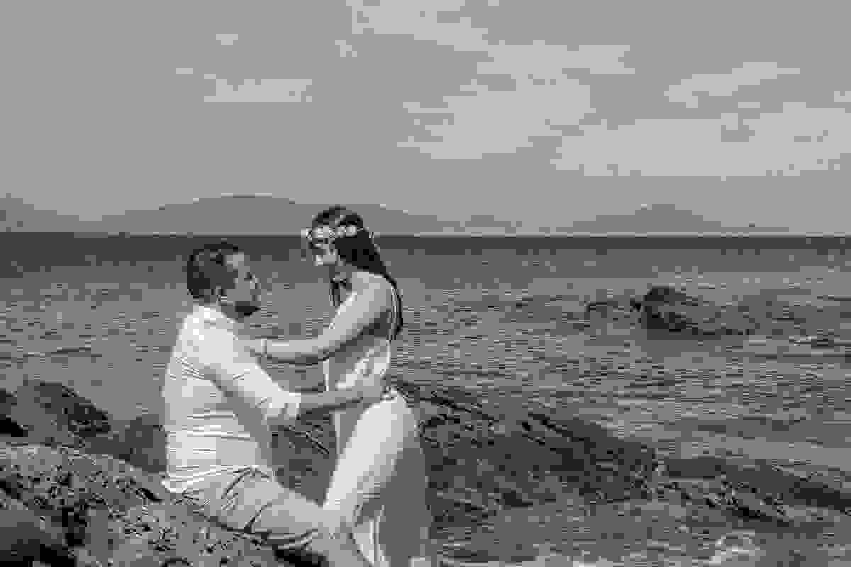 Noivo sentado em uma das pedras e noiva de pé de frente para ele em lindo ensaio pré casamento em cabo frio rio de janeiro