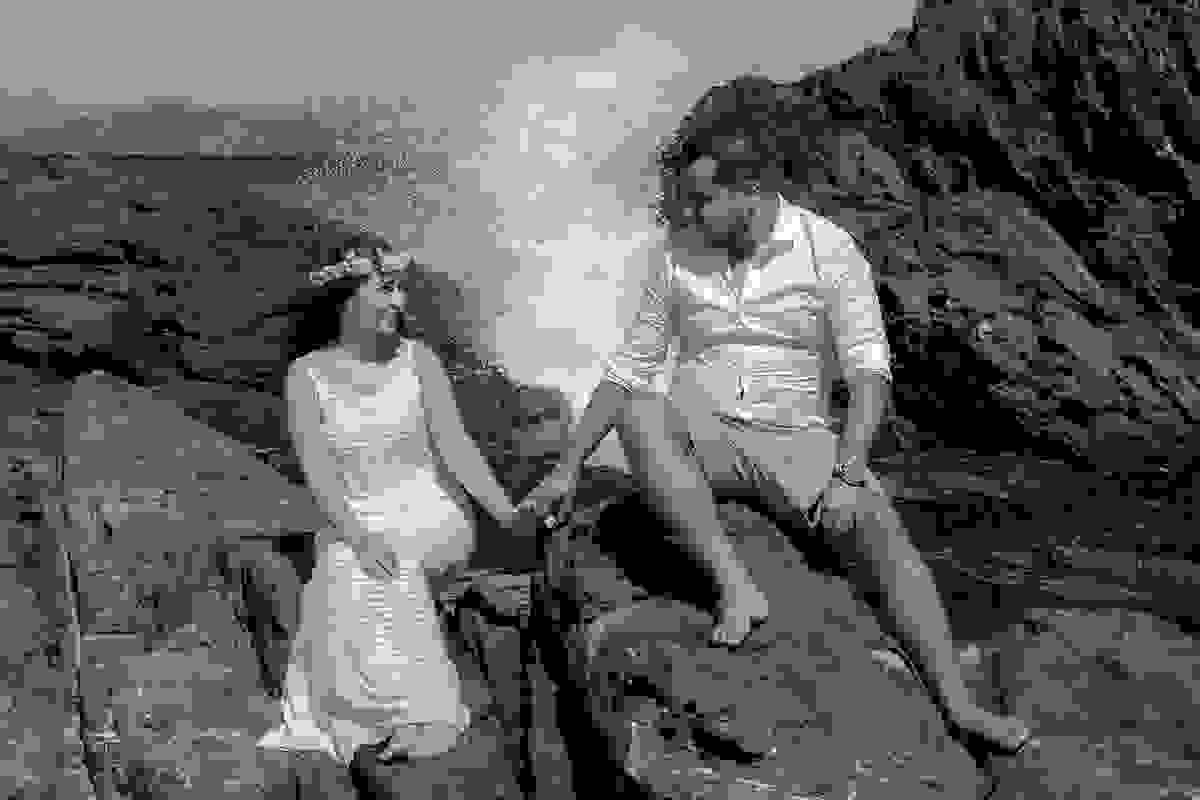 Casal sentado em cima de pedras proximo ao mar com uma onda batendo atraz deles em linda foto de pré casamento na praia de cabo frio rio de janeiro