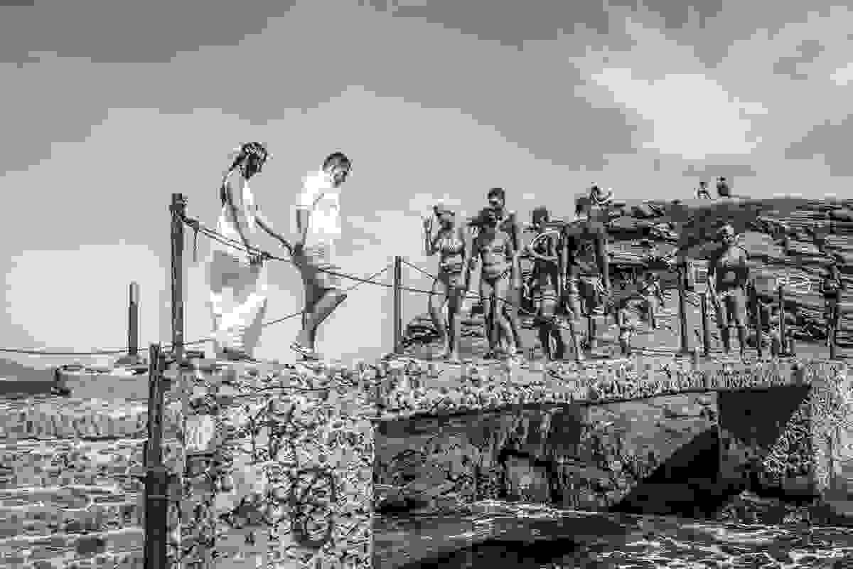 Casal atravesando uma ponte velha com diversos banhistas vindo em sua direção em fotografia de ensaio pré casamento
