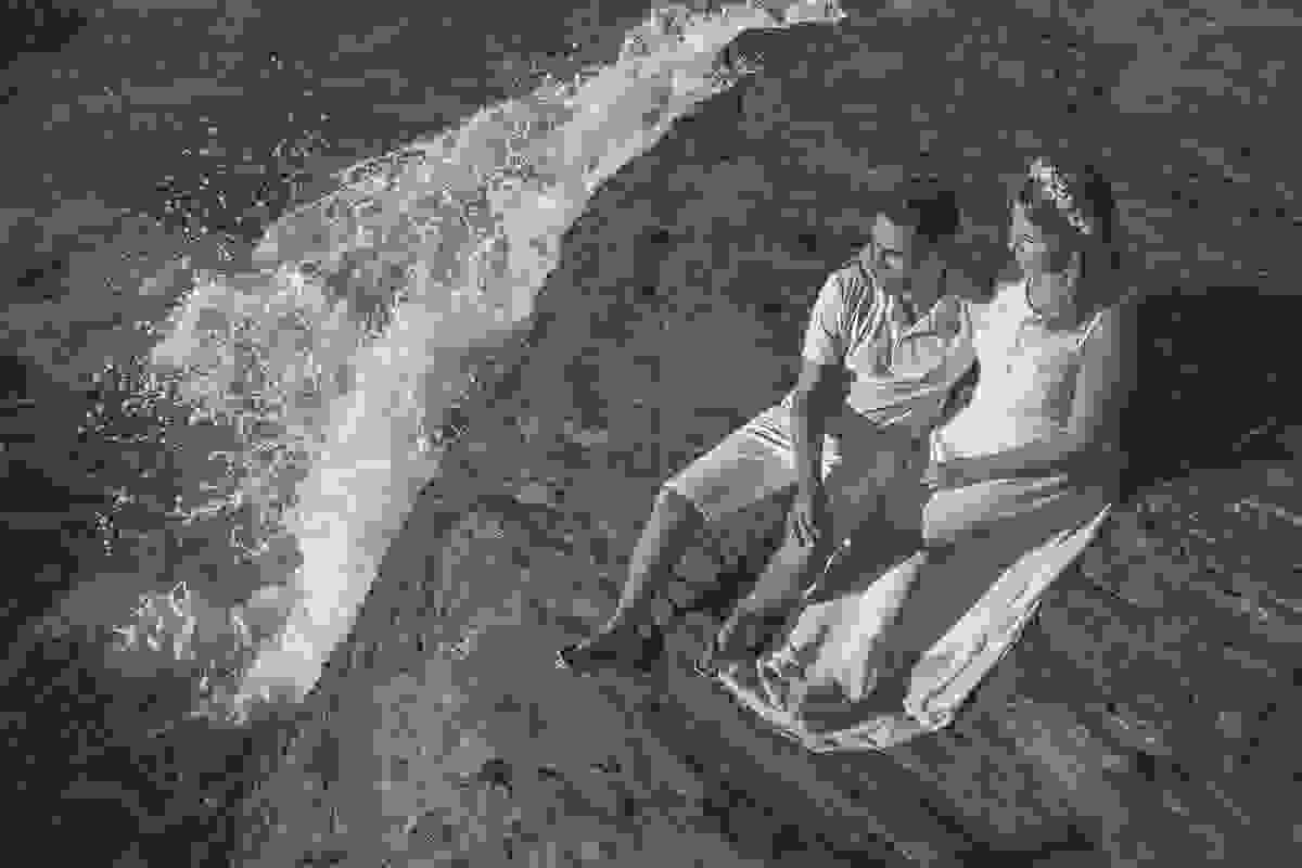Casal sentado em uma pedra com as ondas do mar batendo em ensaio pré casamento na praia