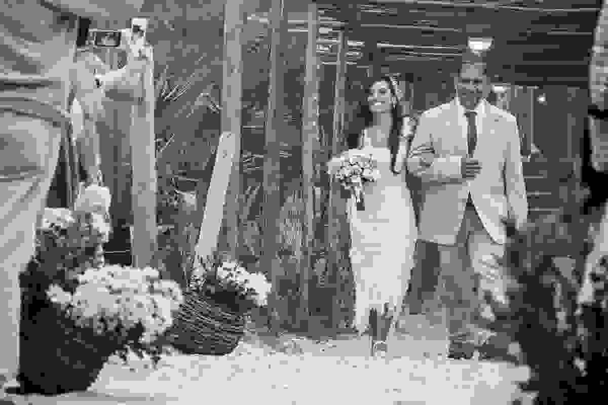 noiva sorrindo com o bouquet em uma das mãos e de braços dados com seu pai caminhando em direção ao noivo