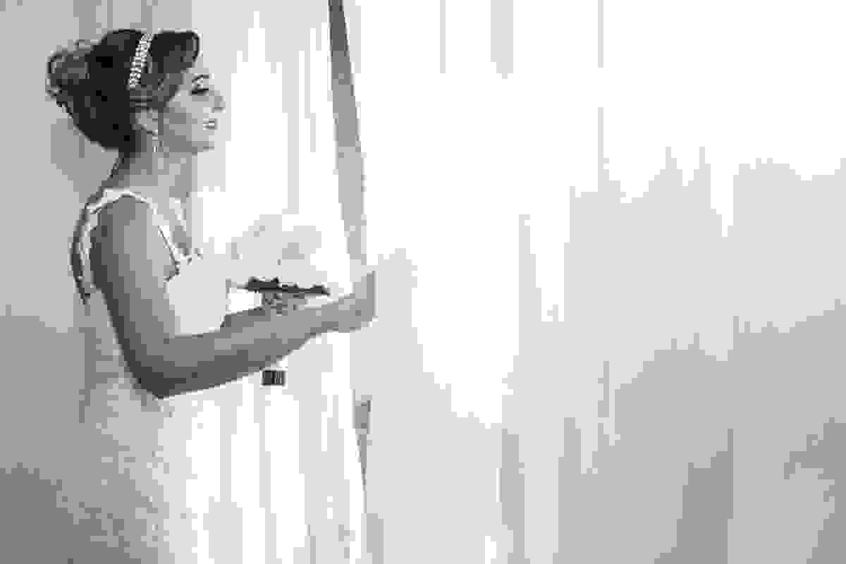 Noiva olha pela janela com bouquet na mão casamento