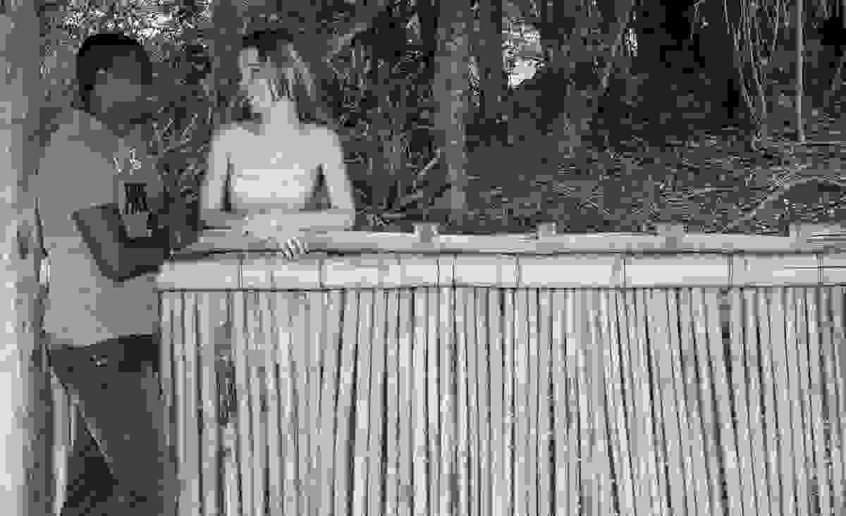 fotografia do casal no ensaio pré casamento ela esta debruçada em um cercado de palha e ele em pé do outro lado olhando para ela