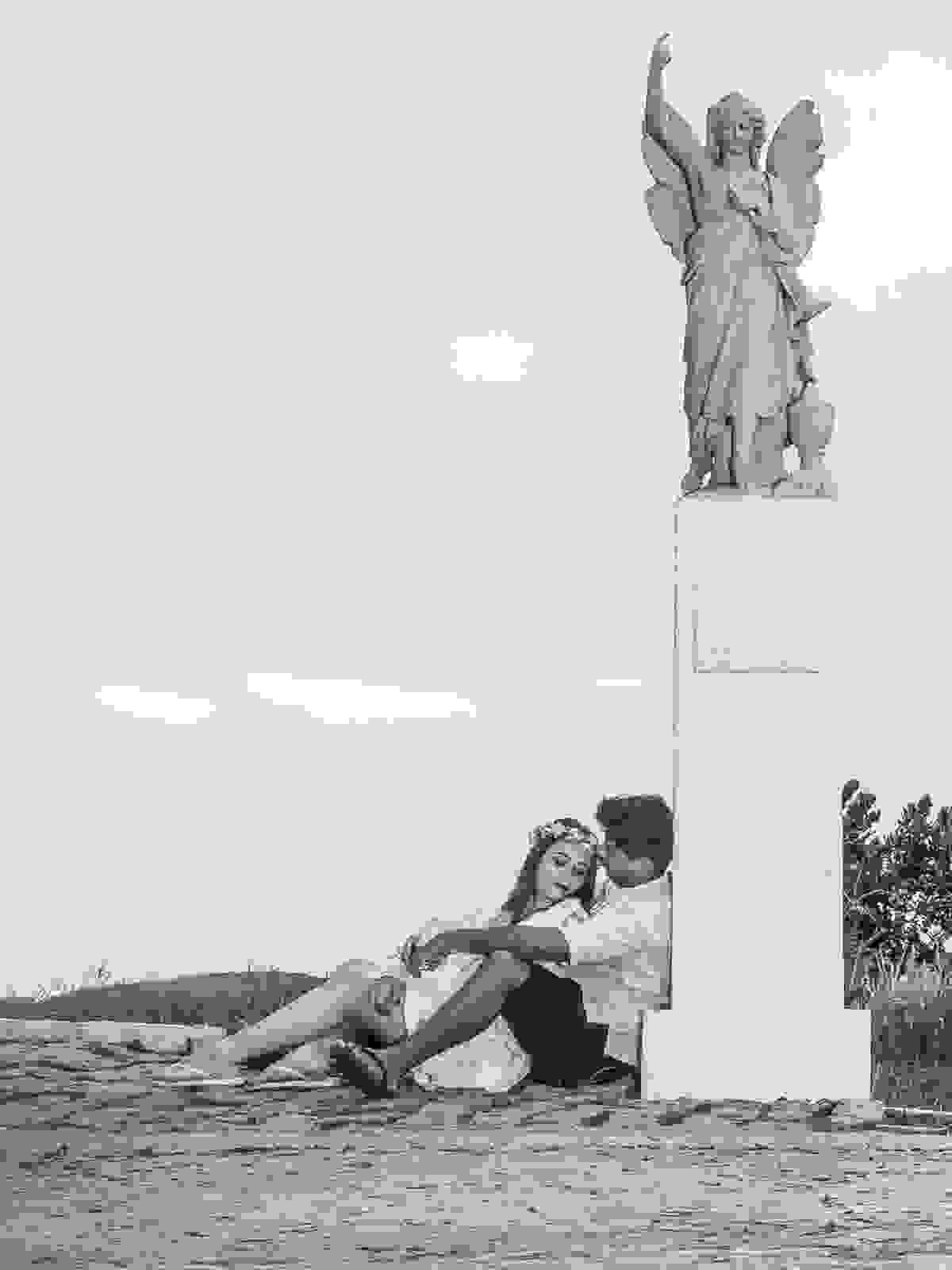 Casal abraçado recostado na base da estátua do anjo em fotografia de pós casamento em armação de búzios Rio de Janeiro