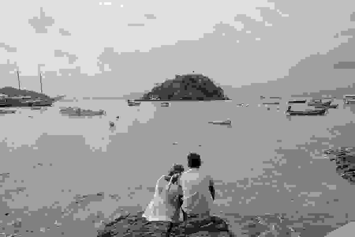Casal viajou para lua de mel em búzios rio de janeiro e registro fotografias como esta que mostra o casal sentado de frente para a praia observando uma ilha ao funco