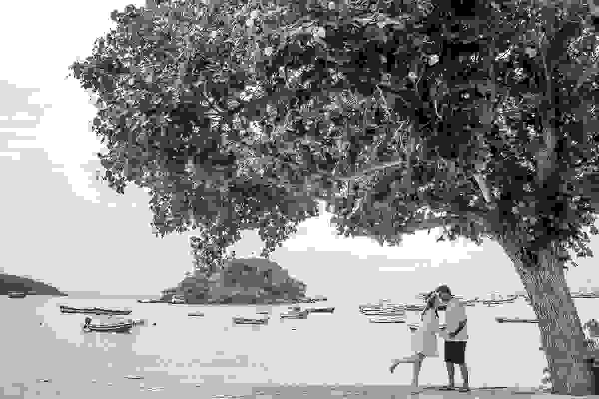 casal e pose embaixo de arvore ela com os pés levantado se inclinando para um beijo nele em fotografia de ensaio pos casamento em armação dos búzios rio de janeiro