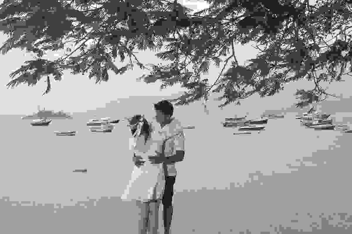 Fotografia de casamento em armação dos búzios mostra casal em ensaio fotográfico abraçados em baixo de uma arvore com o mar ao fundo