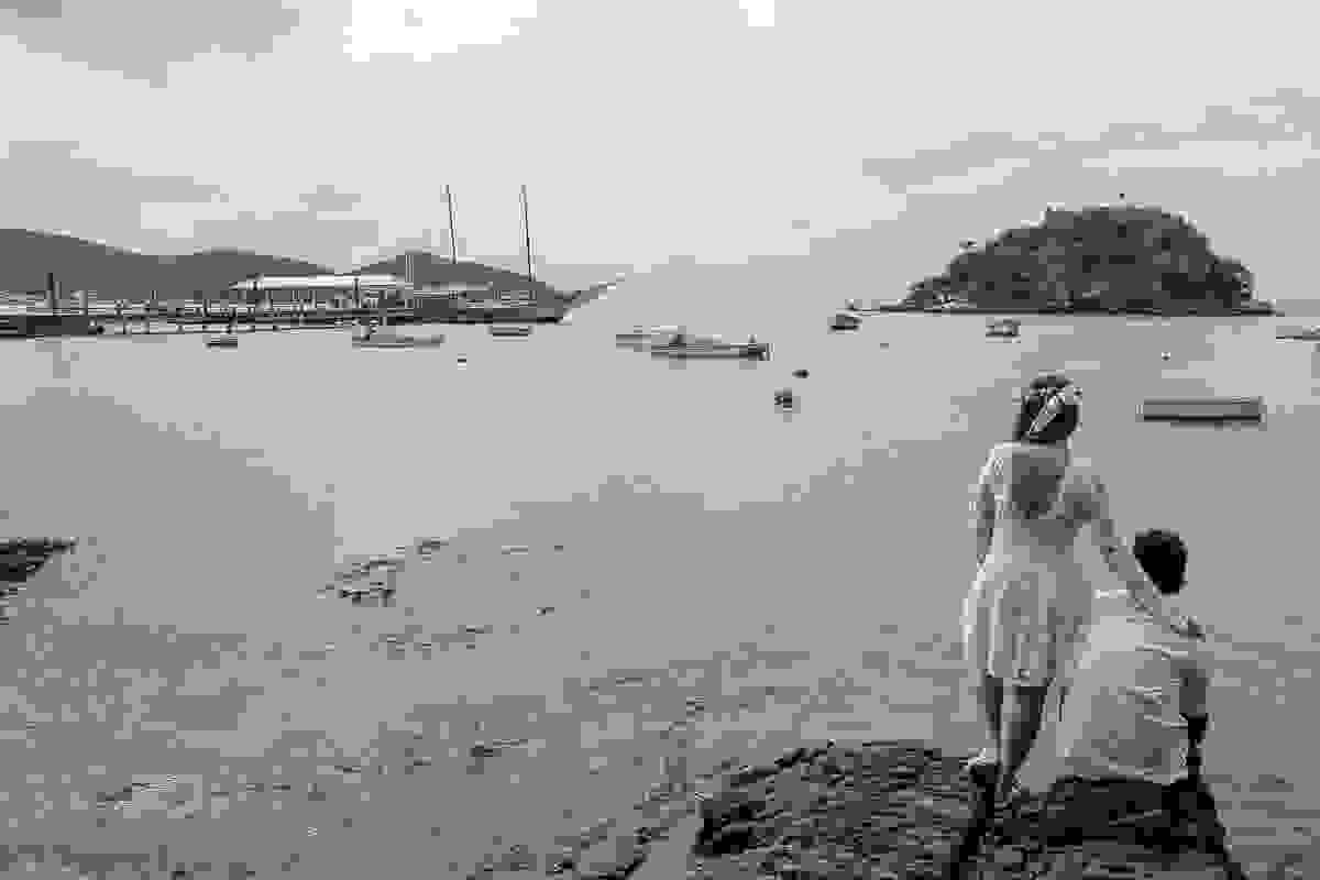 casal ela em pé e ele sentado em uma pedra de frente para a praia da armação em Búzios rio de janeiro no ensaio pré casamento