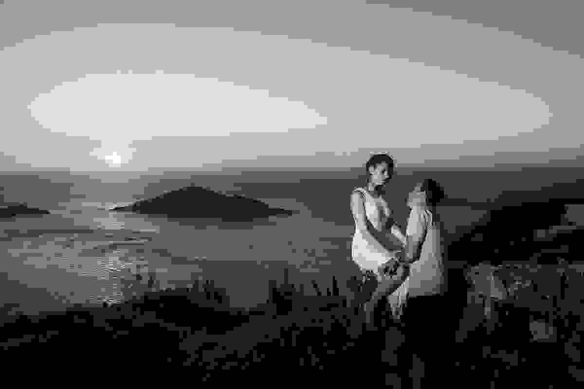 Foto romantica do casal ela sentada em uma pedra e ele olhando para ela e ao fundo o lindo por do sol romantico de arraial do cabo