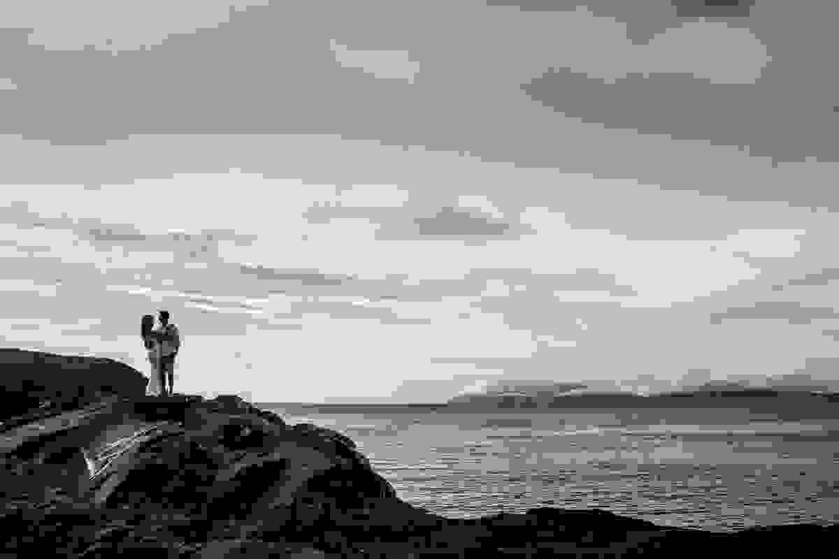Linda fotorgafia em preto em branco com casal de pé sobre uma grande pedra e ao fundo o mar de cabo frio