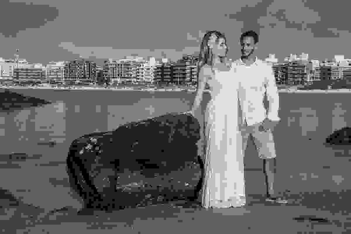 fotografia mostra ao fundo a praia do forte de cabo frio rio de janeiro, casal de pé, ela com a mão encostada em uma pedra