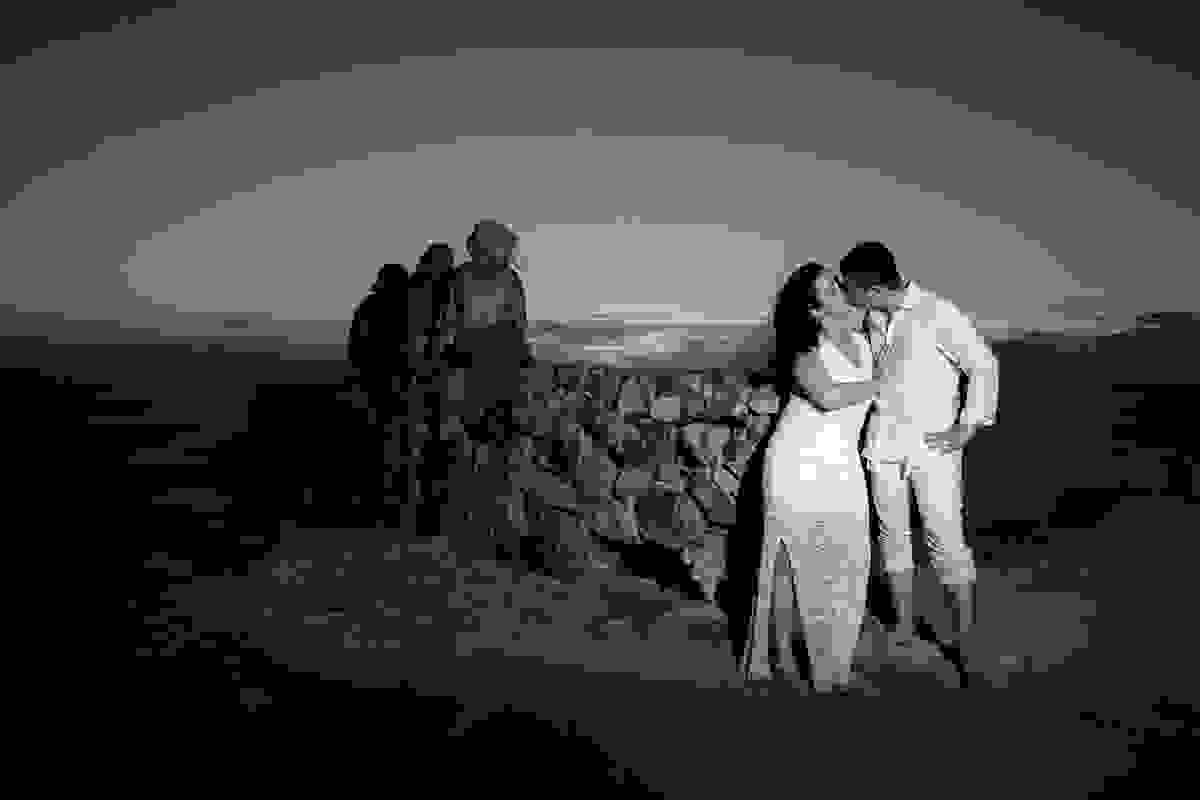 Um beijo apaixonado do casal e ao fundo 03 pessoas sentadas em um muro observando o final do por do sol de arraial do cabo