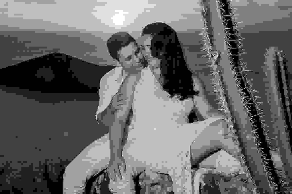 fotografia mostra o início do por do sol em arraial do cabo e os noivos estão sentados em um pequena mureta em uma cena romantica