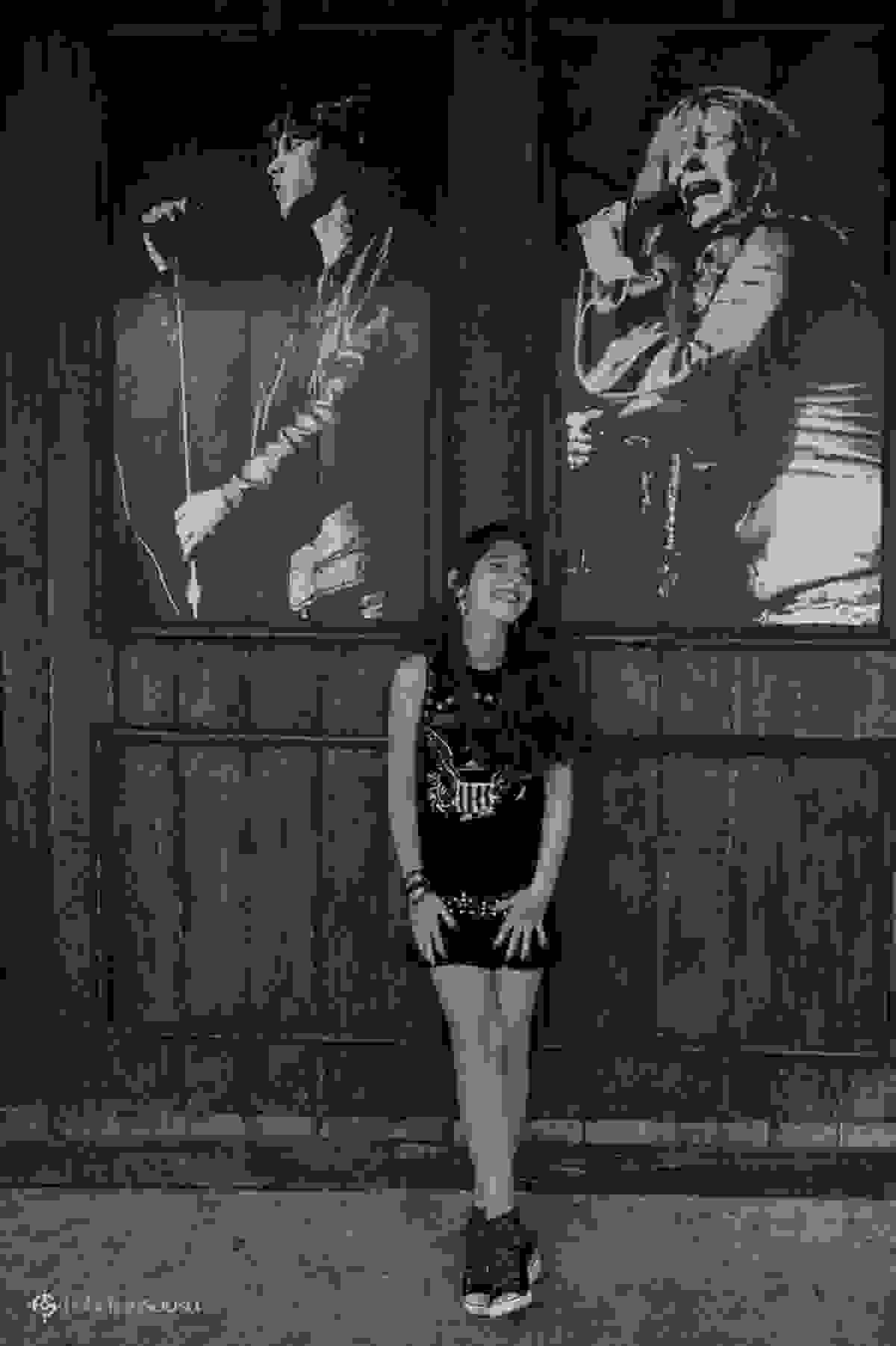 Juliana feliz com roupa preta estilo rock em frente de duas imagens de rockeiros famosos
