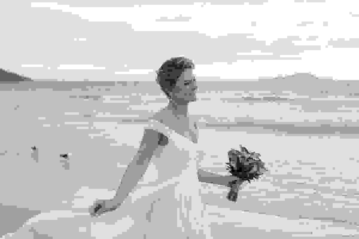 noiva maravilhosa segurando o vestido e o bouquet na beira do mar
